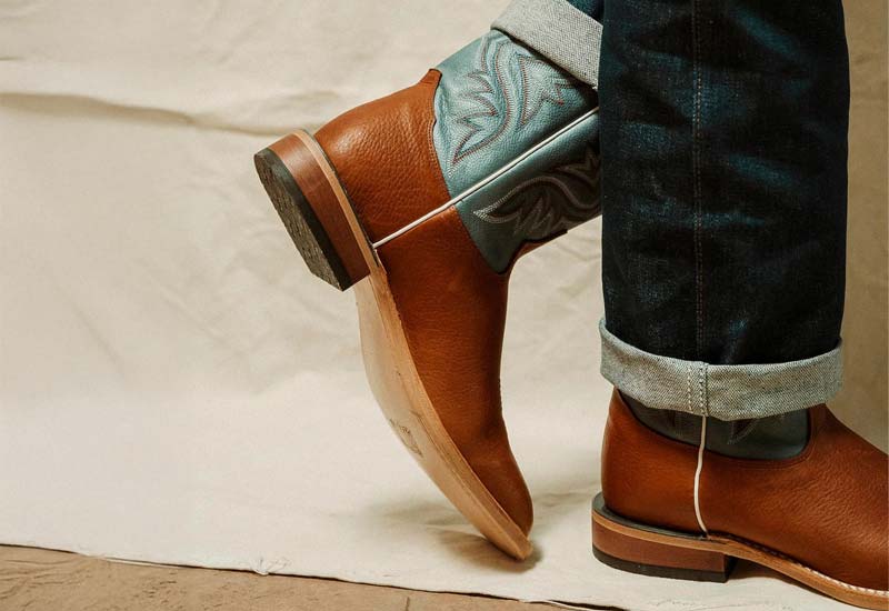 A man wearing a pair of the Avett 11" Western boot in teal and brown.